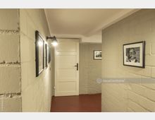 Kleiner Flur mit cremefarben gestrichenen Backsteinwänden und rotem Boden. An den Wänden hängen gerahmte Schwarz-Weiß-Fotografien, und eine Wandlampe beleuchtet den Bereich vor einer weißen Holztür.