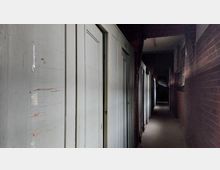 Ein dunkler Korridor mit abgenutzten, weißen Holztüren auf der linken Seite und roten Ziegelwänden auf der rechten Seite. Am Ende des Gangs ist ein schwach beleuchteter Bereich sichtbar.