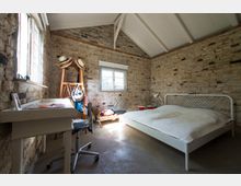 Schlafzimmer mit rustikalem Design, charakterisiert durch freiliegende Ziegelwände und ein weiß gestrichenes Holzgebälk. Der Raum enthält ein schlichtes weißes Bett, einen Schreibtisch mit Drehstuhl, einen Kleiderständer und ein Fenster, das Tageslicht hereinscheinen lässt.