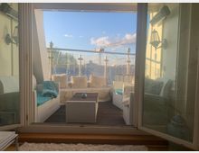 Blick auf eine gemütlich eingerichtete Terrasse mit Holzboden, offenen Glastüren, weißen Rattanmöbeln mit blauen Kissen und dekorativen Kissen aus Kunstfell. Im Hintergrund ein Glasgeländer mit Lichterketten, dahinter blauer Himmel und ein Baukran.