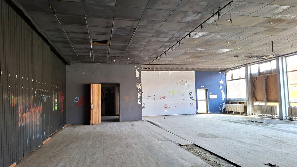 Großer, leerer Innenraum eines verlassenen Gebäudes mit abgehängter Decke, beschädigtem Boden und grauen Wänden mit Graffiti. Rechts verläuft eine Fensterfront mit Heizkörpern darunter, hinten befindet sich eine offene Tür zu einem dunkleren Nebenraum.