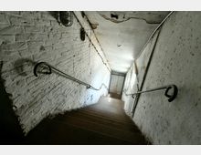 Treppe in einem schmalen, altmodischen Gang mit weißen, teils beschädigten Ziegelwänden, Metallgeländern und spärlicher Beleuchtung. Am unteren Ende des Ganges befindet sich eine geschlossene Holztür.