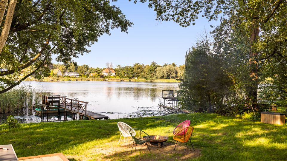 Blick von einer grasbewachsenen Uferwiese auf einen ruhigen See, eingerahmt von großen Bäumen und Schilf, mit zwei kleinen Holzstegen und Sitzgelegenheiten am Wasser. Im Vordergrund stehen drei farbige Gartenstühle um eine Feuerstelle, am gegenüberliegenden Ufer sind Häuser und Bäume zu sehen.