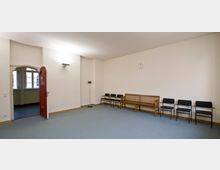 Ein schlichter Warteraum mit hellen Wänden, türkisfarbenem Teppichboden und einer Reihe von Stühlen sowie einer Holzbank an der rechten Wand. Links befindet sich eine offene Tür mit rotem Holzbogen, durch die Fenster im angrenzenden Raum sichtbar sind.