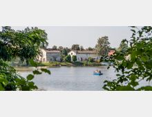 Eine ruhige Seelandschaft mit mehreren Häusern am Ufer, umgeben von Bäumen. Im Vordergrund fährt eine kleine Gruppe von Menschen in einem blauen Ruderboot auf dem See.