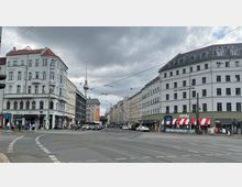 Straßenkreuzung in einer Stadt mit mehrstöckigen Gebäuden im Altbau- und modernen Stil, darunter ein Hotel mit der Aufschrift "The Circus Hotel". Im Hintergrund ist der Berliner Fernsehturm sichtbar, und über der Kreuzung verlaufen Oberleitungen für Straßenbahnen.