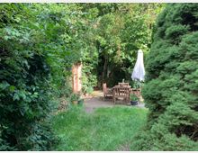 Ein kleiner Garten mit dichtem grünen Bewuchs, einem Holztisch und Stühlen auf einer gepflasterten Fläche. Rechts steht eine große Konifere, und im Hintergrund ist eine Hauswand mit einer Glastür sichtbar.