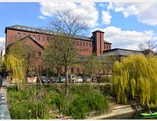 Backsteinbau im Industriedesign mit großen Fenstern und einem Turm, umgeben von Bäumen und Grünflächen. Im Vordergrund fließt ein Kanal mit einer Person in einem grünen Kajak, daneben sitzt jemand auf einer Grasfläche.