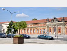 Straßenansicht mit einem historischen Gebäude im Barockstil mit orange-gelber Fassade, Arkadenfenstern und einem reich verzierten Eingang mit Skulpturen. Im Vordergrund sind parkende und fahrende Autos, ein Baum sowie eine bepflanzte Holzkiste.