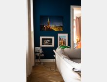 Wohnzimmer mit einer dunkelblauen Wand, an der drei gerahmte Bilder hängen, darunter ein Nachtmotiv mit Kirche und Graffiti. Im Vordergrund ein beige Sofa, ein Holzboden, ein Modell eines Science-Fiction-Roboters auf dem Boden und ein Raum mit Vorhängen im Hintergrund.