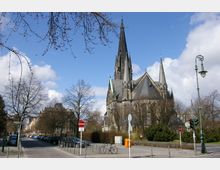Neugotische Kirche mit hohen Türmen und spitzem Hauptturm, umgeben von Bäumen und Straßenlaternen. Im Vordergrund eine Straßenkreuzung mit parkenden Autos, Verkehrsschildern und einem Fahrradständer.