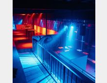 Eine moderne Club- oder Veranstaltungsfläche mit farbiger Beleuchtung in Blau- und Rottönen. Zu sehen sind erhöhte Sitzbereiche, Balkongeländer und ein schlichtes Interieur mit Tischen und hohen Stehtischen.