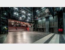Blick auf eine leere Theaterbühne mit schwarzem Boden und technischen Einrichtungen wie Beleuchtungsgerüsten und Vorhangsystemen. Im Hintergrund befinden sich rote Sitzreihen eines Zuschauerraums, umgeben von Vorhängen und Beleuchtungselementen.