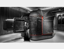 Großer, schwarzer, kuppelartiger Aufbau mit roten Akzenten und handschriftlichen Notizen auf der Oberfläche, ausgestellt in einem Innenraum mit grauen Wänden und Vorhängen. Im Vordergrund ein Schwarz-Weiß-Foto zweier Männer auf einer Stele, im Hintergrund sind weitere Fotografien an der Wand zu sehen.