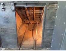 Nahaufnahme eines industriellen Ofens mit geöffneter Stahltür, hergestellt von „Topf & Söhne“. Im Inneren sind Gitterroste und glühende Backsteine zu sehen, mit einem langen Metallwerkzeug davor.