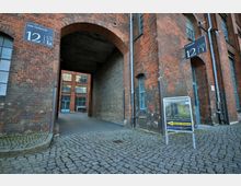 Backstein-Gebäude mit einem großen Torbogen, der zu einem Innenhof führt. Kopfsteinpflaster bedeckt den Boden, und ein Schild weist auf eine Führung durch den "ATG-Tunnel" hin.