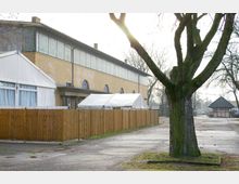 Straßenszene mit einem gelben Backsteingebäude, das große, halbrunde Fenster und ein Fensterband in der oberen Etage aufweist. Im Vordergrund stehen ein Holzzaun, ein weißes Zelt und ein großer Baum, während im Hintergrund kleinere Gebäude und Bäume sichtbar sind.