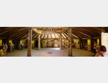 Ein großer, runder Innenraum aus Holz mit sichtbaren Balken und zentralen Holzsäulen stützt ein konisch zulaufendes Dach. Der Raum ist von Vitrinen mit Exponaten und gelblicher Beleuchtung umgeben, während eine Gruppe Menschen entlang der Wände steht.