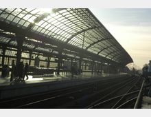 Ein moderner Bahnhof mit großen, gewölbten Glasdächern, die das Bahnsteiggelände überspannen. Im Bild sind mehrere Gleise, beleuchtete Bahnsteige und schemenhafte Personen in einer hellen, sonnendurchfluteten Umgebung zu sehen.