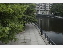 Gepflasterter Gehweg entlang eines Flussufers, gesäumt von Geländern und einer Laterne, mit überhängenden Bäumen links und einem mehrstöckigen Wohngebäude im Hintergrund.