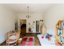 Ein Kinderzimmer mit hellem Holzboden, einem ovalen Holz-Babybett, einem niedrigen weißen Bett mit Kissen und Kuscheltieren sowie einem Bücherregal rechts an der Wand. An der Decke hängt ein schlichter Kronleuchter, und an einer Wand sind Elefanten-Dekorationen und Bilderrahmen angebracht.