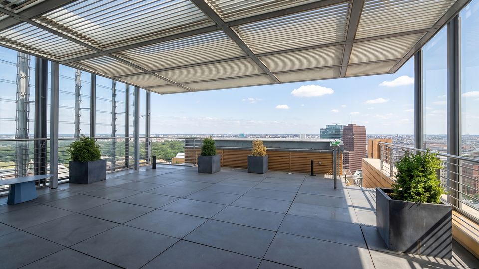 Rooftop observation terrace with large gray floor tiles, metal railings, and a slatted canopy overhead, lined with square planters. A panoramic city skyline and a large green park are visible beyond the glass and open edge.