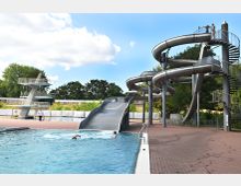 Außenbereich eines Schwimmbads mit einem großen Pool, einer silbernen Wasserrutsche mit Kurven und einer Wendeltreppe sowie einem Sprungturm mit mehreren Plattformen. Im Hintergrund sind Bäume und ein teilweise bewölkter Himmel zu sehen.