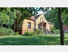 Backsteinhaus mit gelben und roten Ziegeln und einem Satteldach, umgeben von Bäumen und einer Grünfläche im Vordergrund. Das Gebäude verfügt über große Fenster und eine zentrale Treppe, die zu einem überdachten Eingang führt.