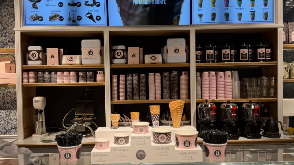 Eine Eisdiele mit einer Glasvitrine, die verschiedene Eissorten präsentiert. Im Hintergrund befinden sich Regale mit Eiswaffeln, Pappbechern und Dekoration sowie digitale Bildschirme, die Eissorten und Getränke bewerben.