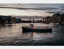 Ein beleuchtetes Boot fährt bei Sonnenuntergang auf einem Fluss, flankiert von modernen Gebäuden. Im Hintergrund erheben sich die Oberbaumbrücke und der Berliner Fernsehturm, während das Schild der „Universal“-Halle rechts gut sichtbar ist.