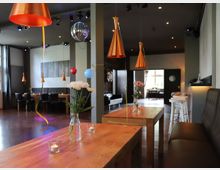 Ein moderner Veranstaltungsraum mit Holz-Esstischen, braunen Polsterbänken und orangefarbenen Hängelampen. Im Raum befinden sich Dekorationen wie Blumen in Glasvasen, hängende Luftballons und eine Discokugel an der Decke, mit einem Fenster im Hintergrund, das Tageslicht einlässt.