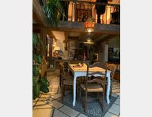 Gemütlicher Innenraum eines rustikalen Hauses mit sichtbaren Holzbalken und einer Galerie im Obergeschoss mit einem schmiedeeisernen Geländer. Im Vordergrund steht ein Esstisch mit hölzernen Stühlen auf gefliesten Böden, dekoriert mit Pflanzen, orientalischen Lampen und ländlichen Wohnaccessoires.