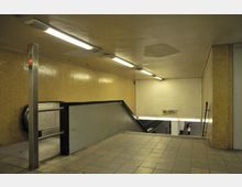 Unterirdischer U-Bahnhof mit gelb gekachelten Wänden, einer Rolltreppe und einem Treppenabgang. Oberhalb sind beleuchtete Schilder und Leuchtstofflampen an der Decke zu sehen.