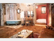 Rustikales Loft mit freigelegten Backsteinwänden, einer freistehenden Badewanne mit Duschvorhang, einem Holztisch mit Spiegel, und einem erhöhten Schlafbereich mit roter Wand und Vorhang. Der Raum hat Holzdielenboden, Vintage-Möbel und dekorative Teppiche.