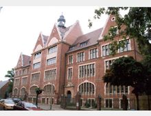 Rotes Backsteingebäude im Stil der Neogotik oder Neorenaissance, mit spitzgiebligen Dachaufbauten, einem kleinen Turm in der Mitte und großen, bogenförmigen Fenstern. Vor dem Gebäude befinden sich ein Zaun, ein Tor und vereinzelt Bäume, sowie geparkte Autos an der Straße.