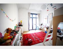 Helles Kinderzimmer mit weiß gestrichenen Backsteinwänden und einer großen Glastür mit Vorhängen, die zu einem Balkon führt. Das Zimmer ist mit Regalen voller Spielzeug, einem roten Teppich mit Mustern, einem Hochbett und einem Schreibtisch ausgestattet.
