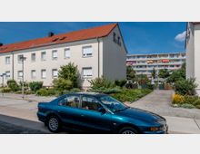 Wohnstraße mit einem zweigeschossigen Mehrfamilienhaus mit weißer Fassade und rotem Satteldach im Vordergrund. Im Hintergrund ein mehrstöckiges Wohnhaus mit bunten Balkonen; davor ein grüner Vorgarten und ein geparkter, dunkelgrüner PKW auf der Straße.