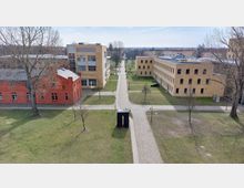 Luftaufnahme eines Campusgeländes mit modernen und historischen Gebäuden. Links steht ein rotes Backsteingebäude, rechts und zentral befinden sich gelbe Neubauten, dazwischen verlaufen gepflasterte Wege und Grünflächen mit vereinzelt stehenden Bäumen.