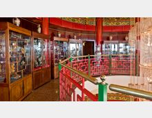 Innenansicht eines Museums mit asiatischer Gestaltung, roten Holzsäulen, verzierten Geländern in Rot und Grün sowie Vitrinen mit Glasfronten, die Kunstwerke und Vasen zeigen. Ein Kronleuchter aus Kristall hängt rechts im Bild.