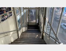 Treppenabgang in einem überdachten, verglasten Bereich eines Bahnhofs mit Metallgeländern auf beiden Seiten. Durch die Fenster sind Teile einer Straße sowie parkende Autos sichtbar.