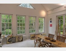 Heller Atelierraum mit großen weißen Sprossenfenstern, die den Blick auf einen grünen Garten freigeben. Im Vordergrund stehen ein Holztisch und Stühle, an den Wänden sind kleine Gemälde aufgehängt, und Staffeleien sowie Malutensilien sind im Raum verteilt.