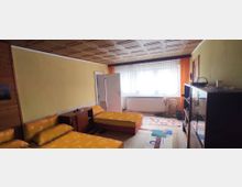 Ein einfach eingerichtetes Schlafzimmer mit zwei Einzelbetten in gelber Bettwäsche, einer Kommode, einem großen Fenster mit weißen Vorhängen und orangefarbenen Seitenvorhängen. Rechts stehen eine dunkle Vitrine und eine Zimmerpflanze, die Fußböden sind mit bunten Teppichen bedeckt.