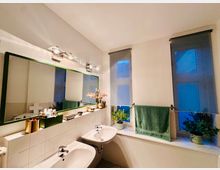 Modernes Badezimmer mit zwei weißen Waschbecken, einem großen rechteckigen Spiegel mit Beleuchtung und mehreren dekorativen Elementen wie Pflanzen und Handtüchern. Drei Fenster mit Rollos lassen gedämpftes Licht herein, und eine kleine Ablagefläche unter dem Spiegel bietet Platz für Pflegeprodukte.