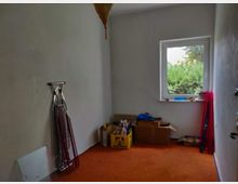 Kleiner, leerer Raum mit orangefarbenem Teppichboden und weiß gestrichenen Wänden. An der linken Wand lehnen ein Bügelbrett und weitere Gegenstände, während vor einem Fenster Kartons, Haushaltsgegenstände und rote Vorhangstangen stehen; durch das Fenster ist grünes Gebüsch sichtbar.