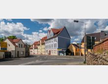 Straßenszene in einem Wohnviertel mit mehrfarbigen Gebäuden im klassischen Stil, darunter ein auffälliges blaues Haus mit roten Dachziegeln. Im Vordergrund befindet sich eine asphaltierte Kreuzung mit Straßenschildern und einer Straßenlaterne, während eine Backsteinmauer rechts den Gehweg begrenzt.
