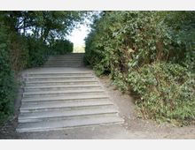 Stein-Treppe in einem parkähnlichen Bereich, umgeben von dichtem grünen Gebüsch und Bäumen, die Schatten spenden. Die Treppe führt leicht nach oben und ist von einem unbefestigten Weg umgeben.