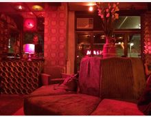 Ein gemütliches Café-Interieur mit warmem, rotem Licht, gepolsterten Sofas, einem dekorativen Regal aus runden Fächern und Blumen in einer Vase. Große Fenster im Hintergrund bieten einen Blick nach draußen, wo beleuchtete Schriftzüge sichtbar sind.