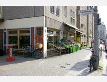 Straßenszene mit einem kleinen Lebensmittelgeschäft an der Ecke eines Wohngebäudes. Auf dem Bürgersteig stehen Obst- und Gemüsestände mit grünem Tuch bedeckt, daneben Fahrräder, ein Coca-Cola-Stehtisch und eine Straßenlaterne mit einem Straßenschild „Auguststraße“.