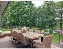 Holzterrasse mit einem großen Esstisch aus Glas und acht geflochtenen Stühlen, umgeben von einer Glasbrüstung. Im Hintergrund ein Garten mit grüner Wiese, Bäumen, einem Spielbereich und einem See.