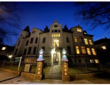 Eine historische Villa bei Nacht, mit einer kunstvollen Fassade und zahlreichen Fenstern, die beleuchtet sind. Im Vordergrund befindet sich ein Tor mit zwei gemauerten Pfeilern, die mit kleinen Dächern verziert sind, und ein schmiedeeiserner Zaun umgibt das Grundstück.